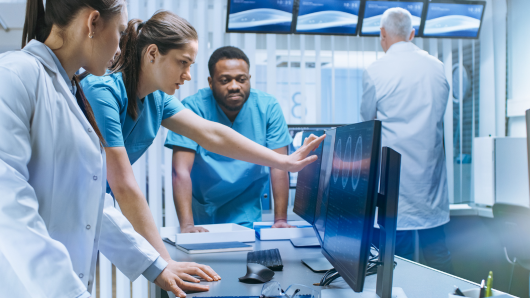 Woman in scrubs pointing to screen with medical results while two other medical professional look on