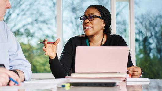 Female executive sits at a table behind a computer and speaks to colleagues.