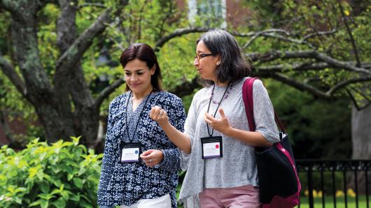 Two female executives wearing badges talk while walking on the HBS campus